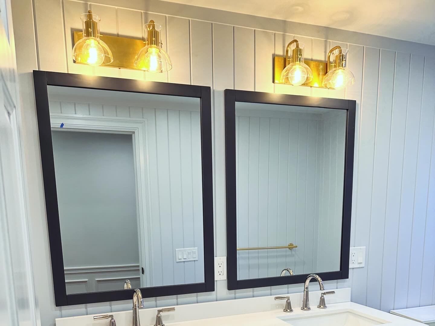 Two black-framed bathroom mirrors above a white double vanity with gold wall sconces.