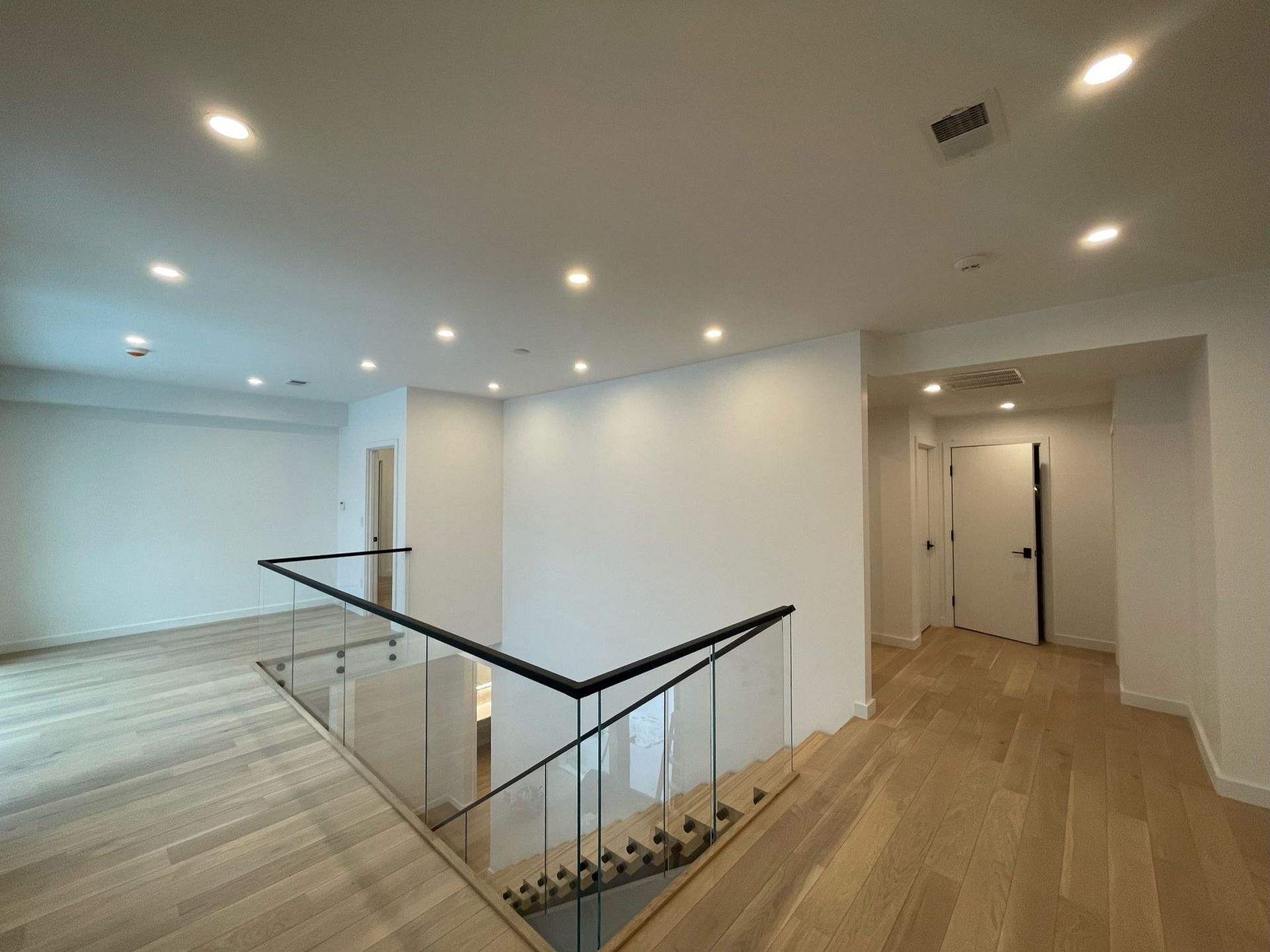 Modern empty home interior with wood floors, recessed lights, and a glass-railed staircase.