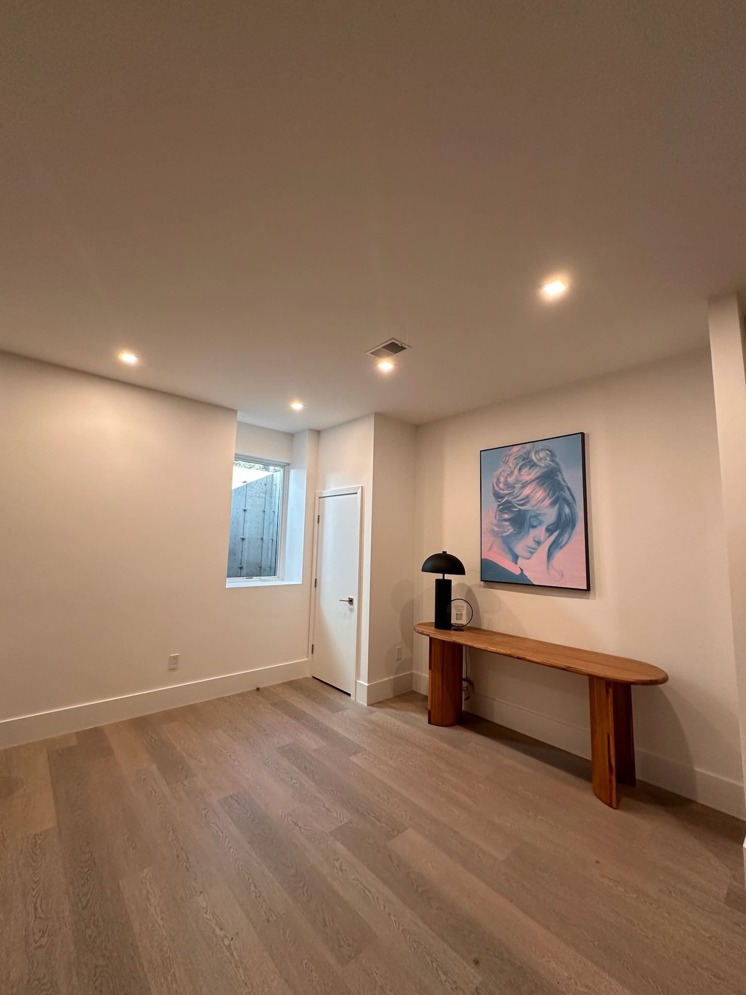 Empty modern room with wood floors, recessed lights, a window, and a console table beneath wall art.