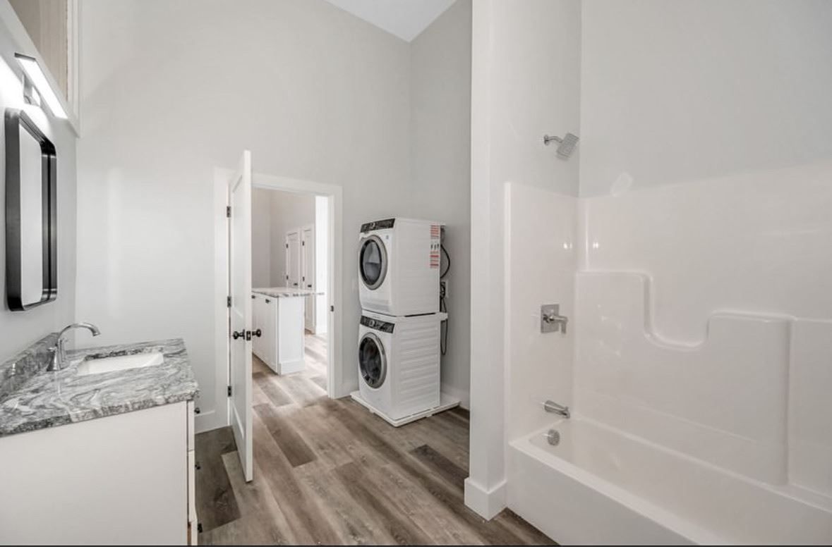 Bright laundry room with stacked washer and dryer, white walls, wood-look floor, and a bathtub on the right