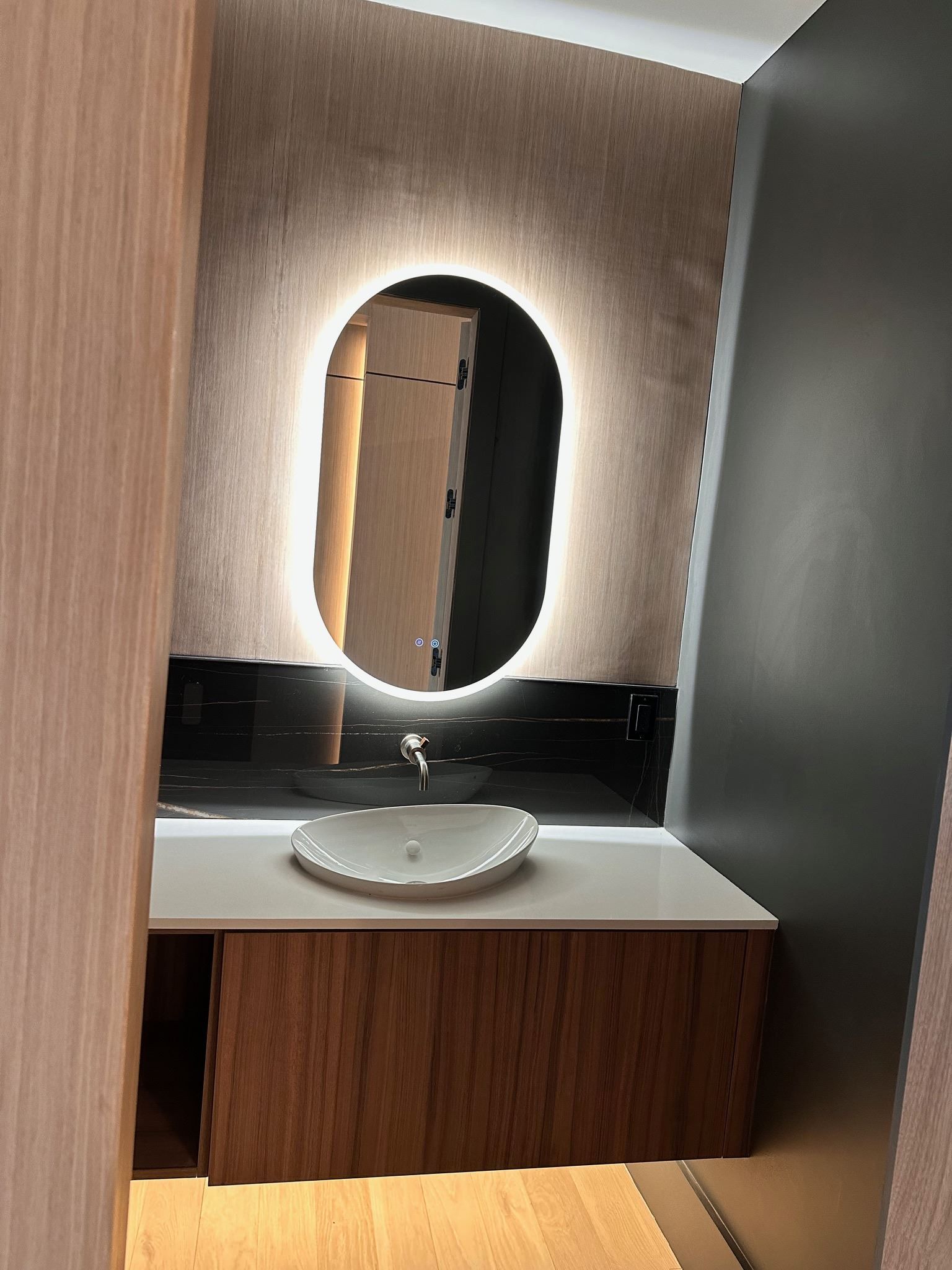 Modern bathroom vanity with oval backlit mirror, white sink, wood cabinetry, and dark wall accents