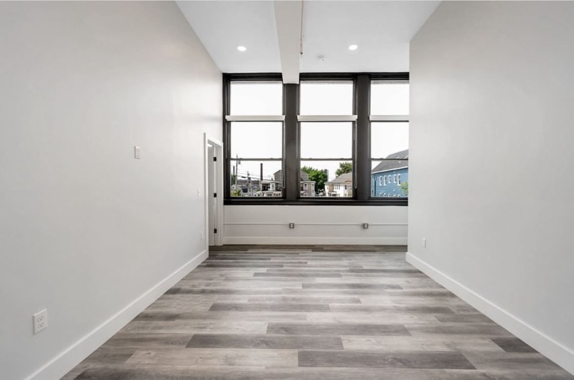 Empty modern room with white walls, gray wood floor, and three windows overlooking city buildings.