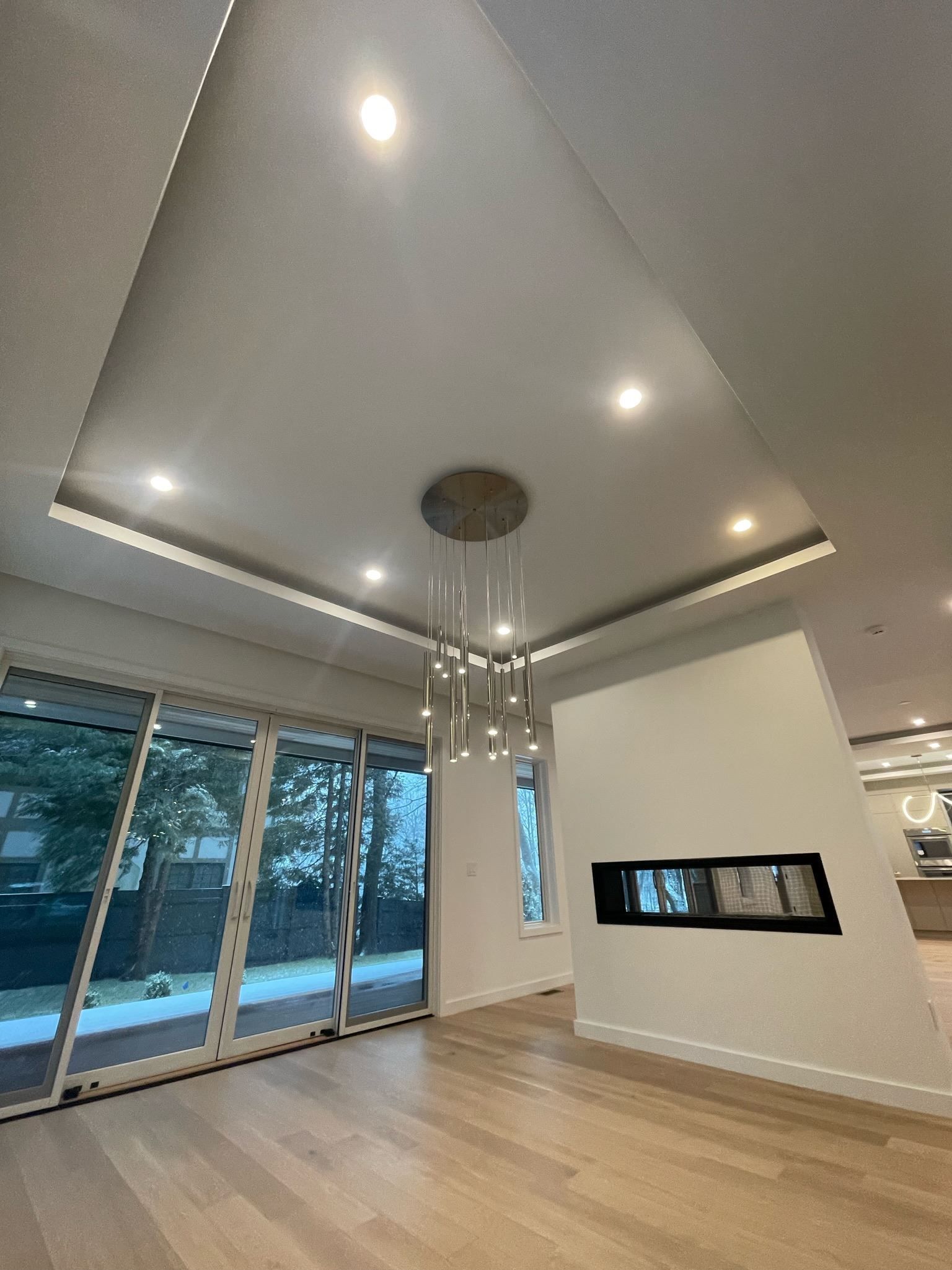 Empty modern living room with recessed lights, wood floor, large windows, and a hanging chandelier.