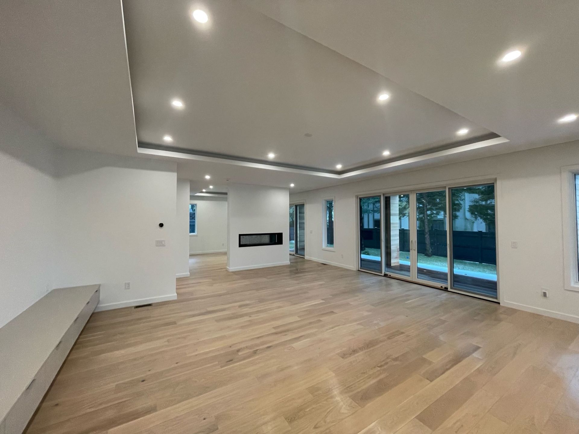 Empty modern living room with light wood floors, recessed lighting, fireplace, and large glass doors to a patio