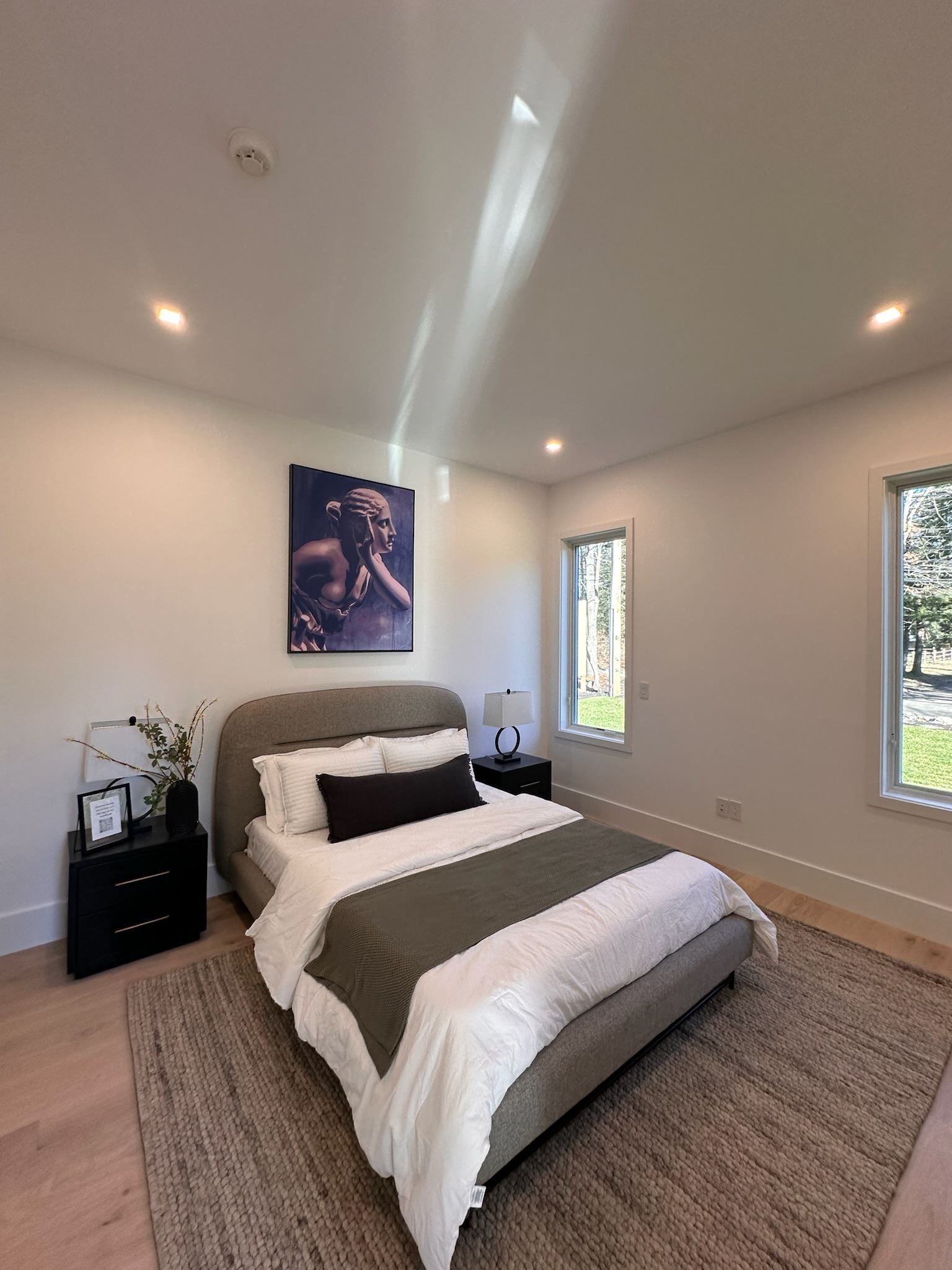 Modern bedroom with a neatly made bed, bedside tables, framed wall art, and bright windows.