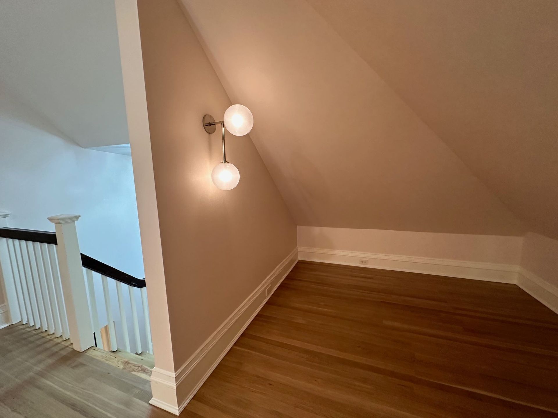 Empty stair landing with sloped beige wall, white trim, and hardwood floor beside a staircase.