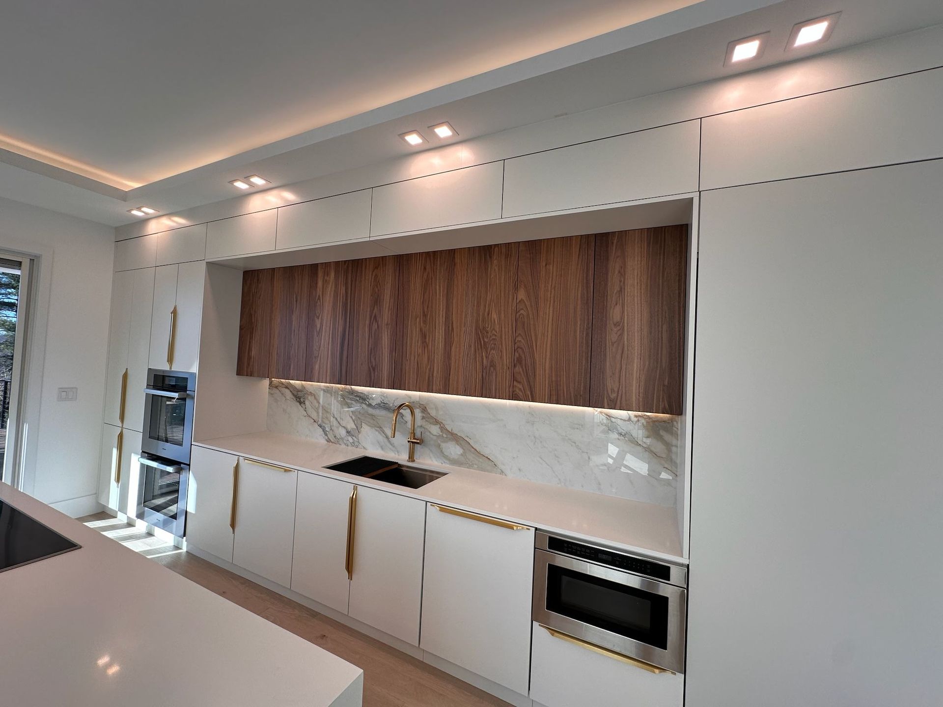 Modern white kitchen with wood cabinets, marble backsplash, built-in oven, and ceiling spotlights