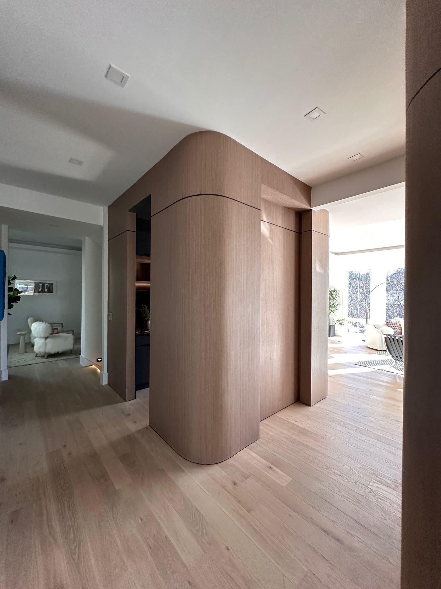 Modern hallway with curved wood-paneled partition, light wood floors, and bright windows at the end