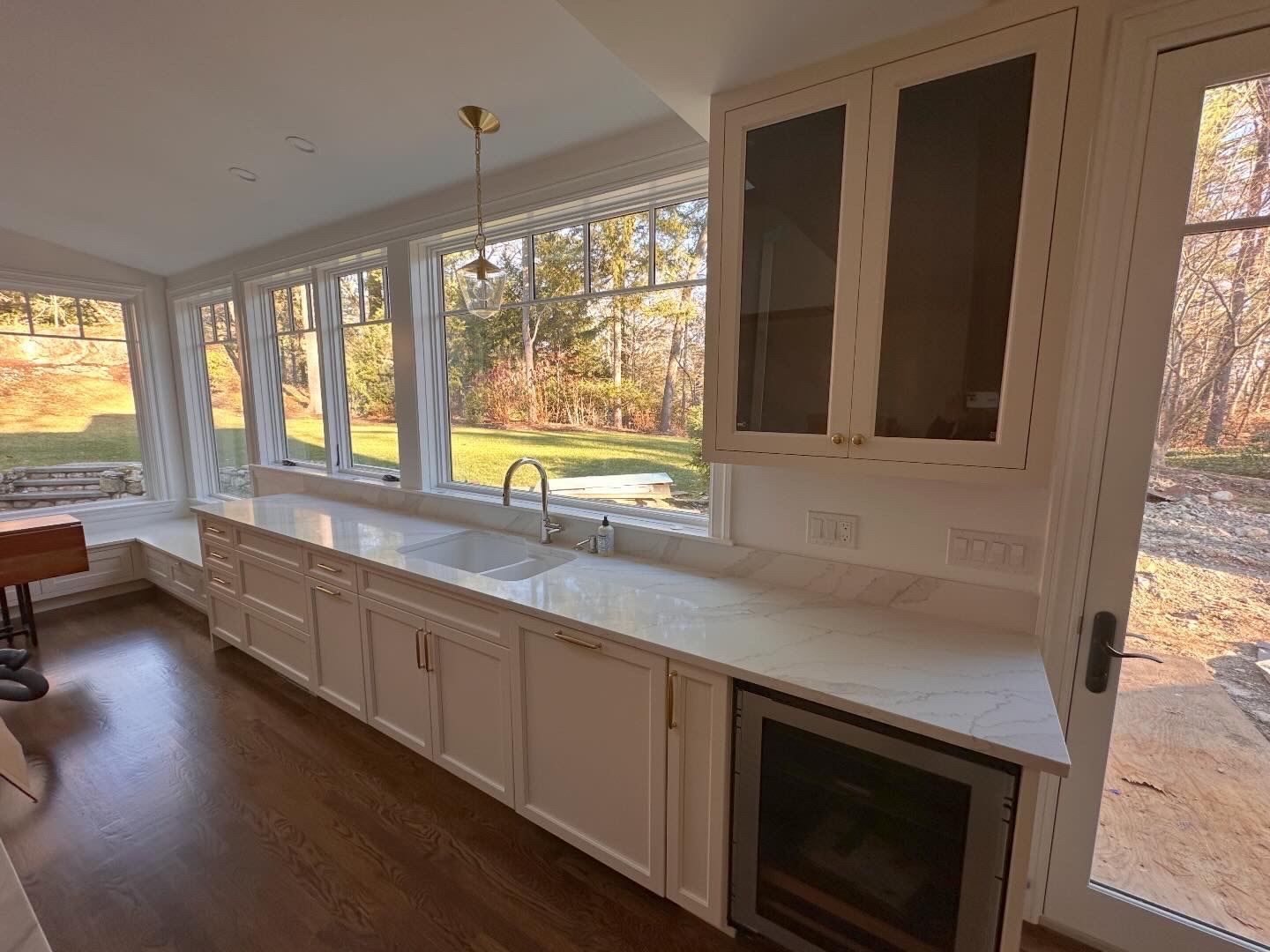 Bright sunroom with long white cabinets, large windows, and dark wood floors overlooking trees outside