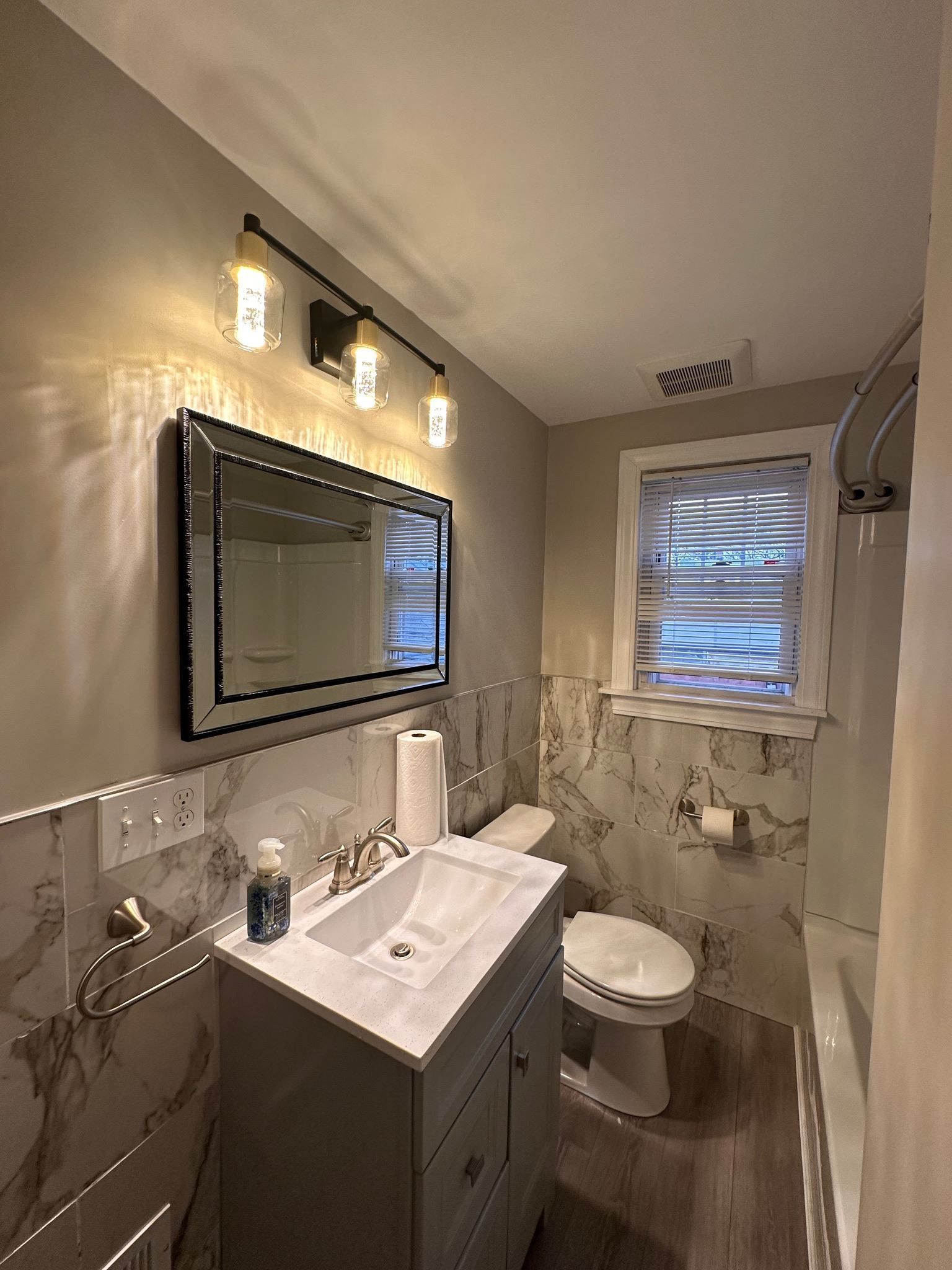 Small beige bathroom with vanity, mirror, toilet, shower, and two windows covered with plastic.