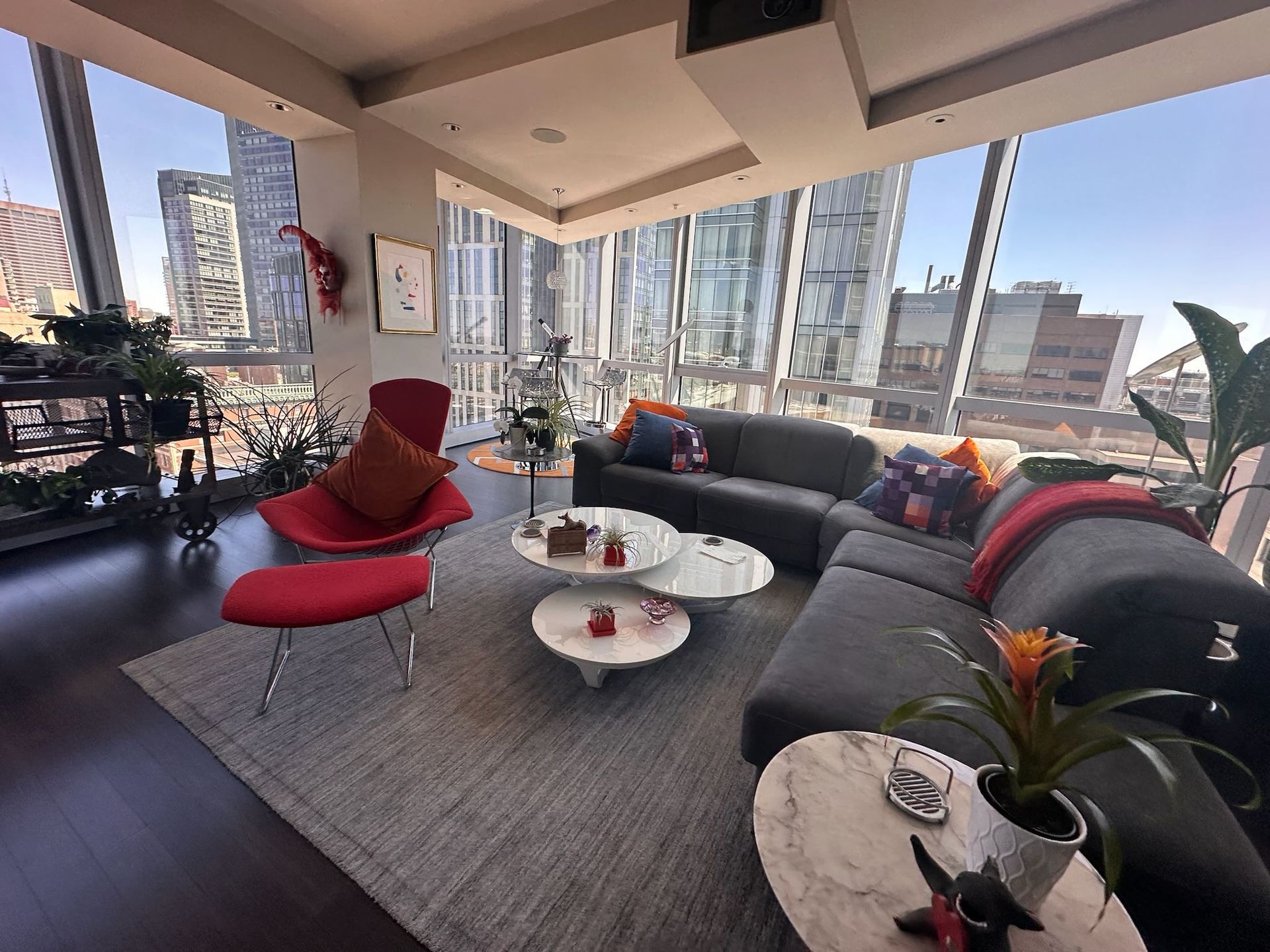 Modern living room with gray sectional, red chair, round tables, and floor-to-ceiling windows overlooking the city