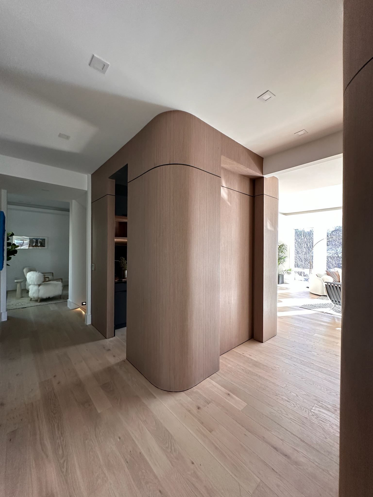 Modern interior hallway with light wood floors and curved wood wall paneling, opening to a bright room.