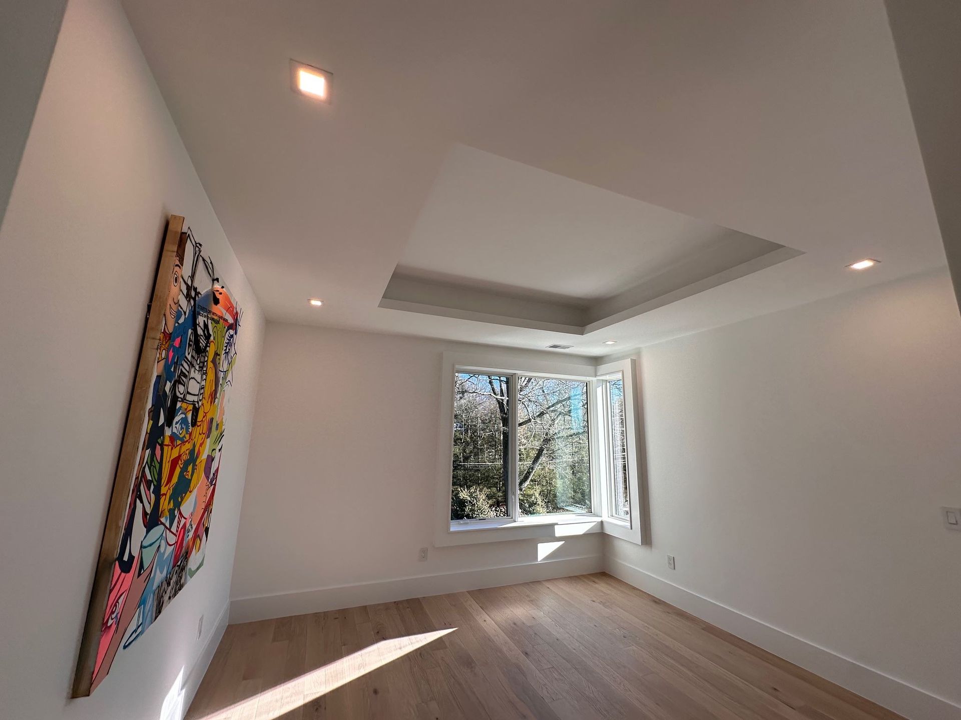 Empty modern room with white walls, wood floor, recessed ceiling lights, and a colorful abstract painting by a window.