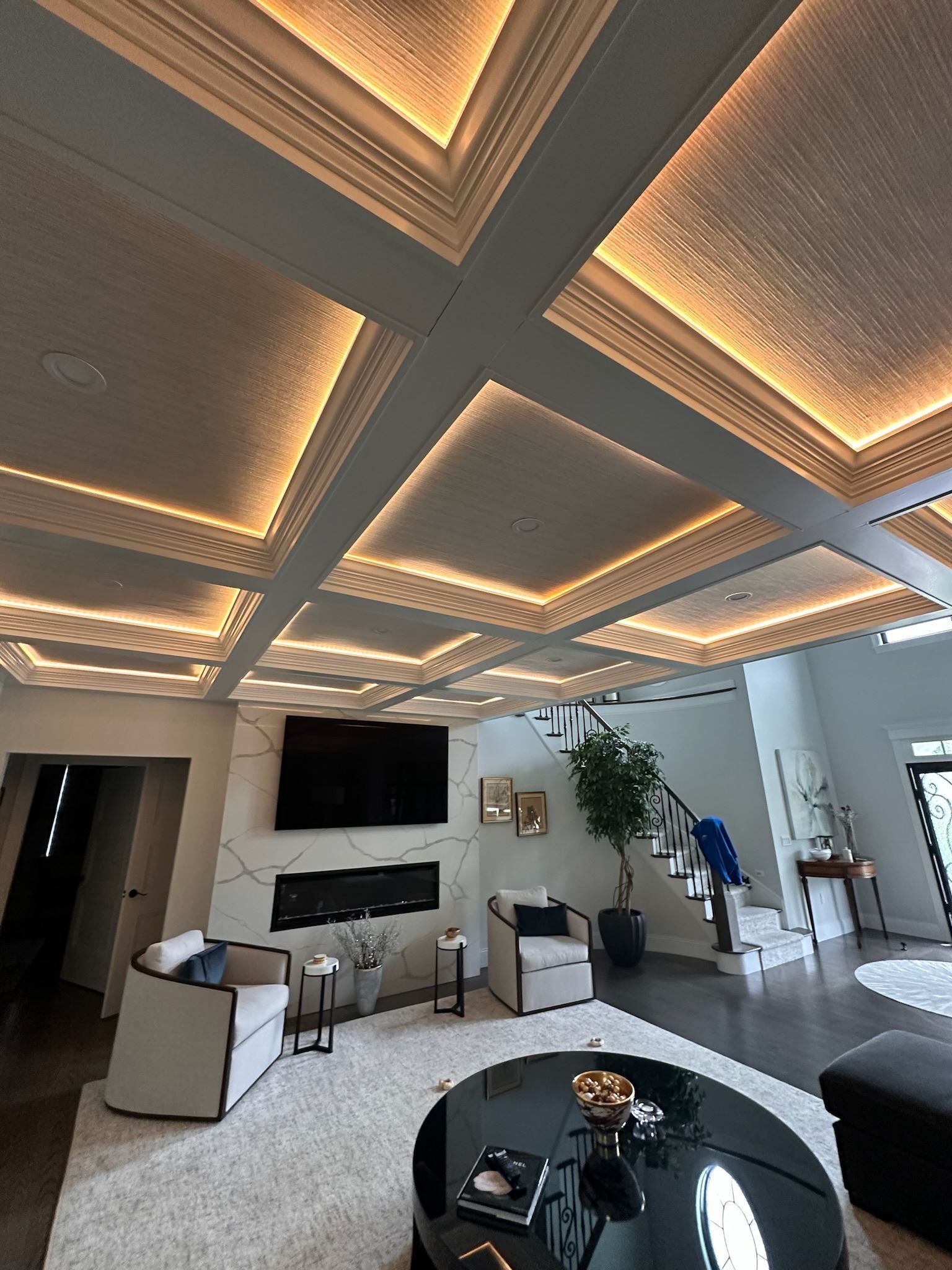 Modern living room with illuminated tray ceiling, TV, sofas, coffee table, and staircase.