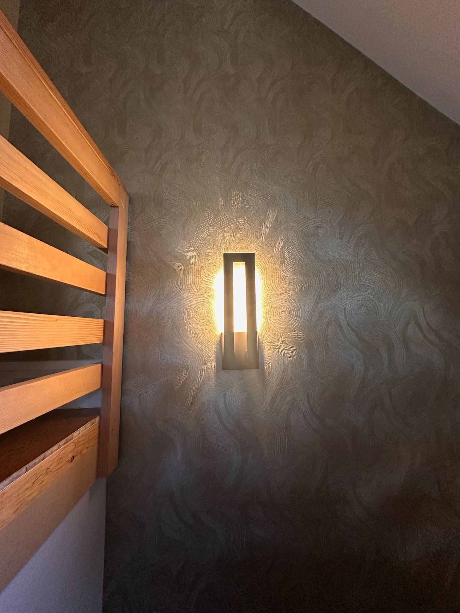 Hallway wall with a glowing sconce beside a wooden staircase railing, lit in warm tones