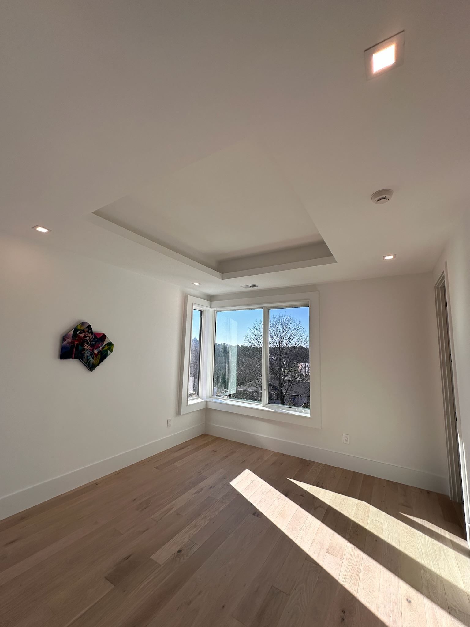 Empty sunlit room with hardwood floors, white walls, and a large window.
