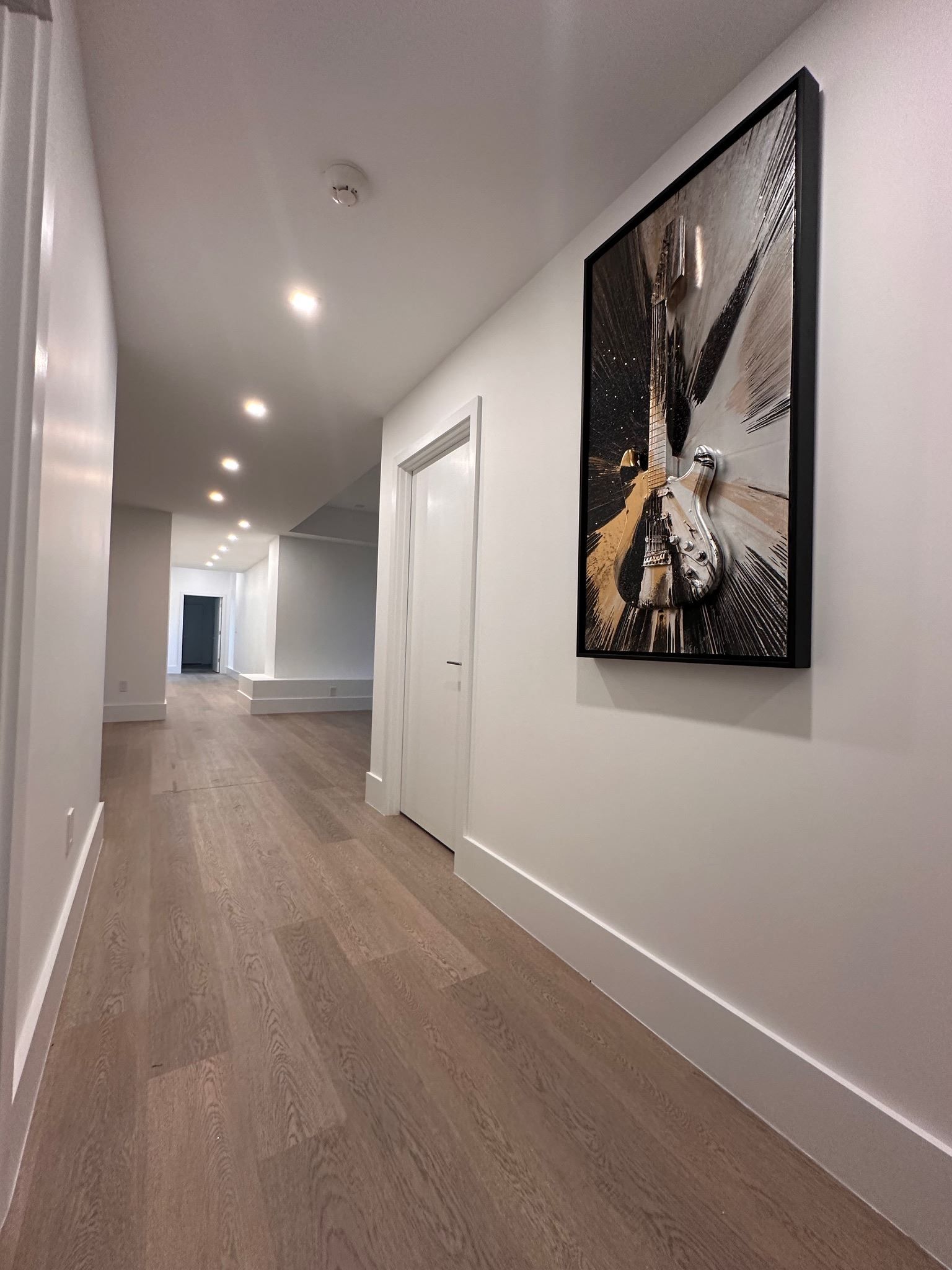 Long modern hallway with wood floors, white walls, recessed lights, and framed artwork.
