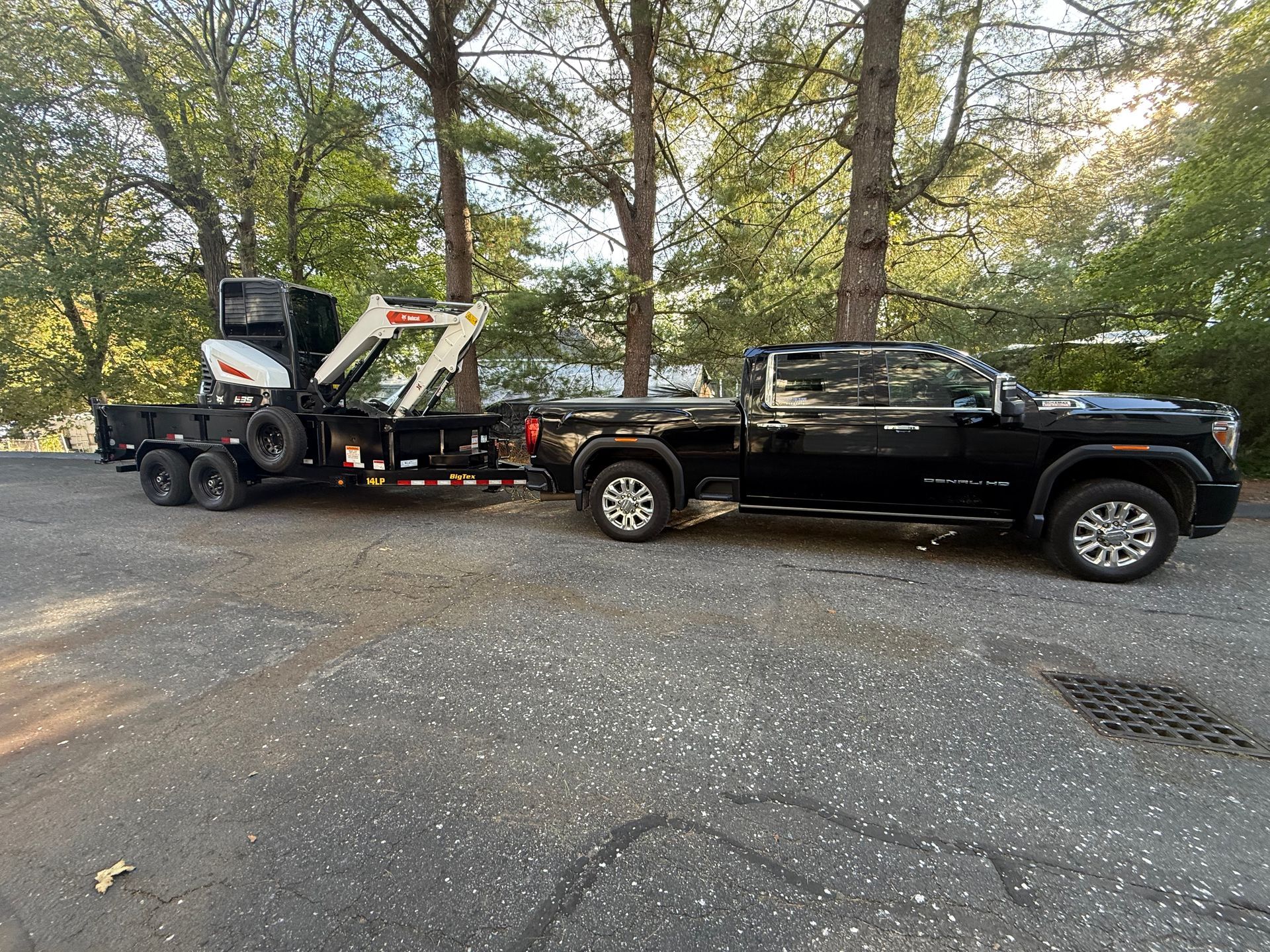 Black pickup towing a trailer with a skid steer on a gravel lot near trees