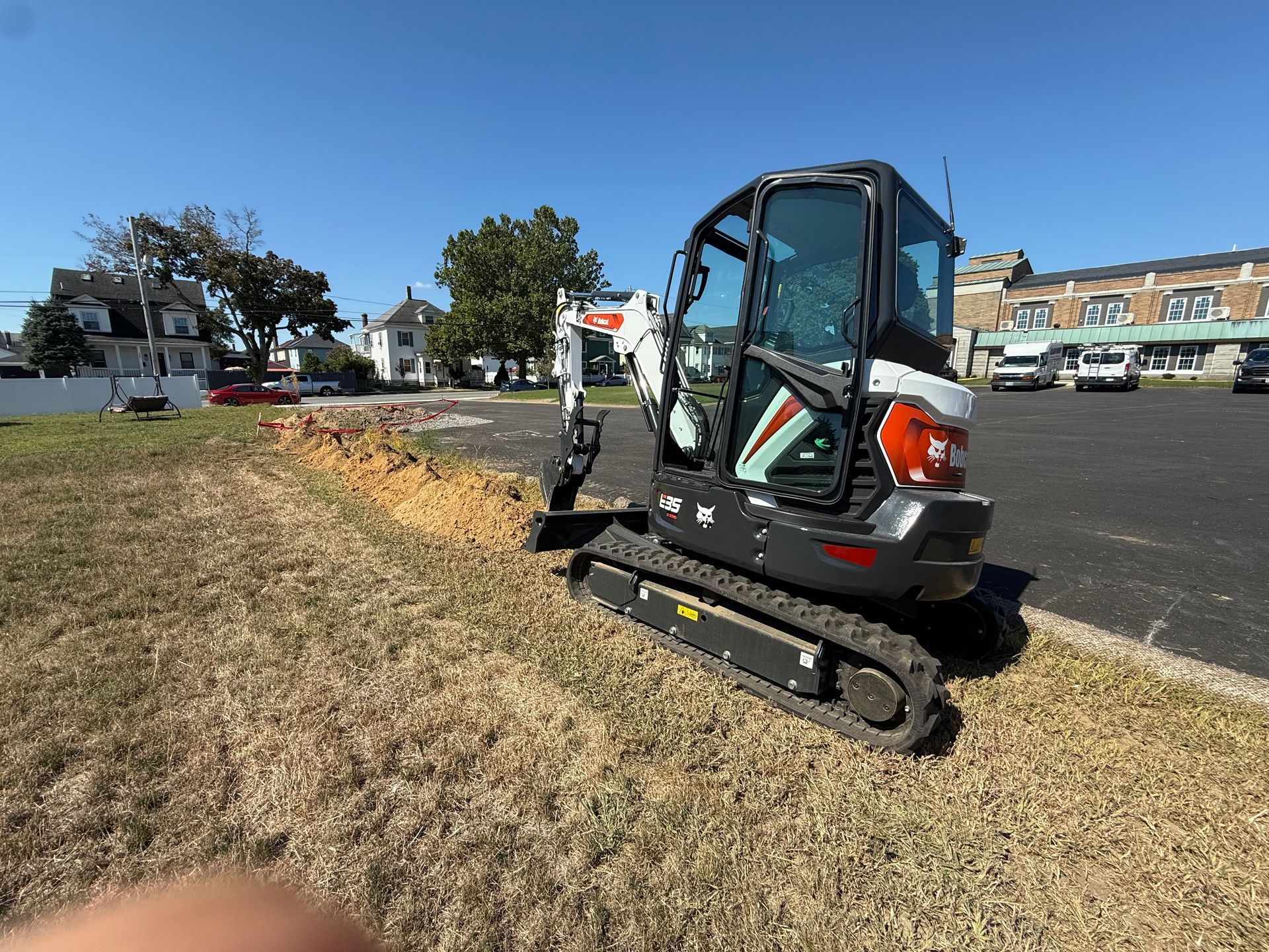 Mini excavator on tracks beside a grassy slope in a parking lot, with houses and trees in the background