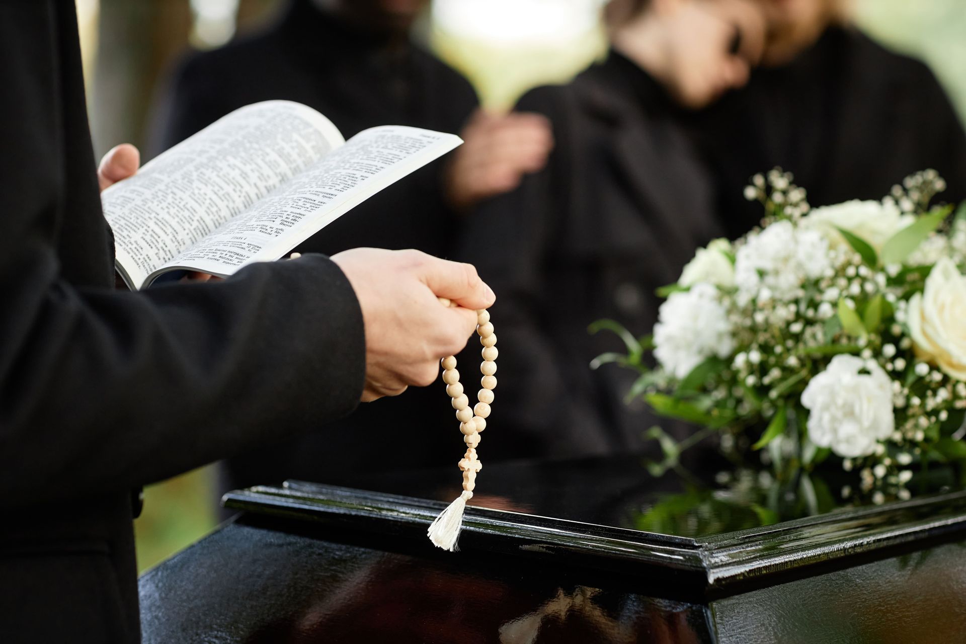 Close up of person in black praying at outdoor funeral and holding Bible with rosary