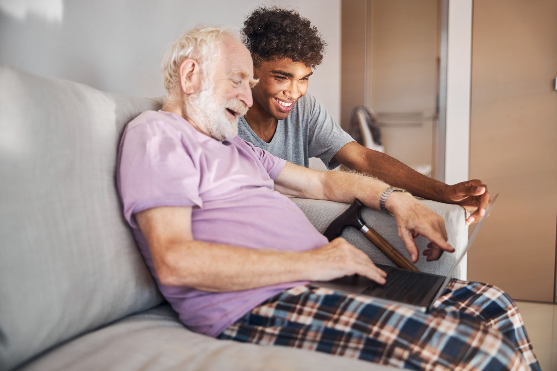 Man and older adult seated on a couch, looking at a laptop. The older adult smiles and points.