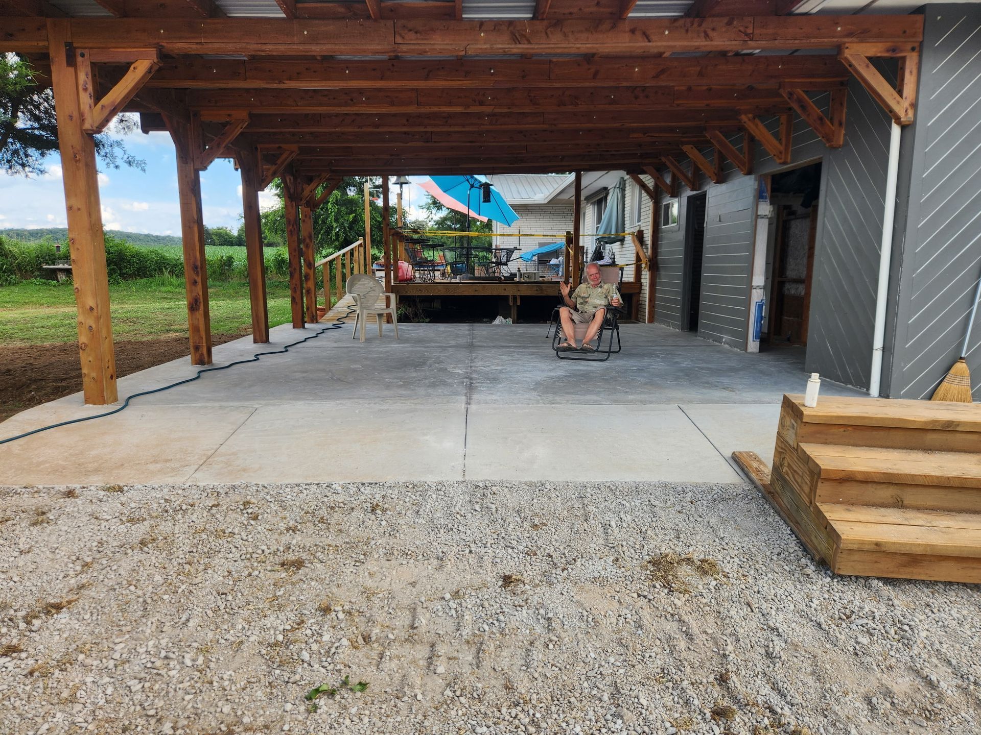 Covered outdoor patio with wooden beams, concrete floor, a grill, and seating by a grassy field.