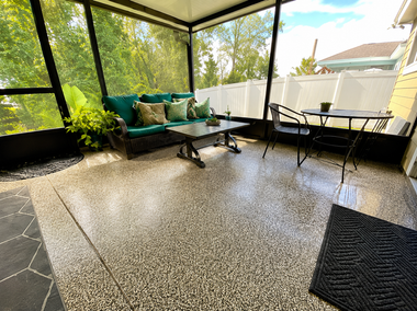 Covered patio with wicker seating, glass table, and speckled tile floor overlooking a backyard.