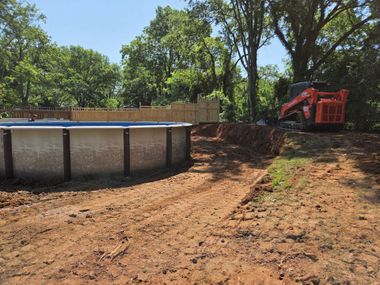 Above-ground pool beside a dirt yard, with a red excavator and trees in the background.