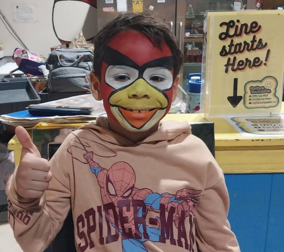Child in a Spider-Man hoodie and bird mask gives a thumbs-up in a classroom or office setting.