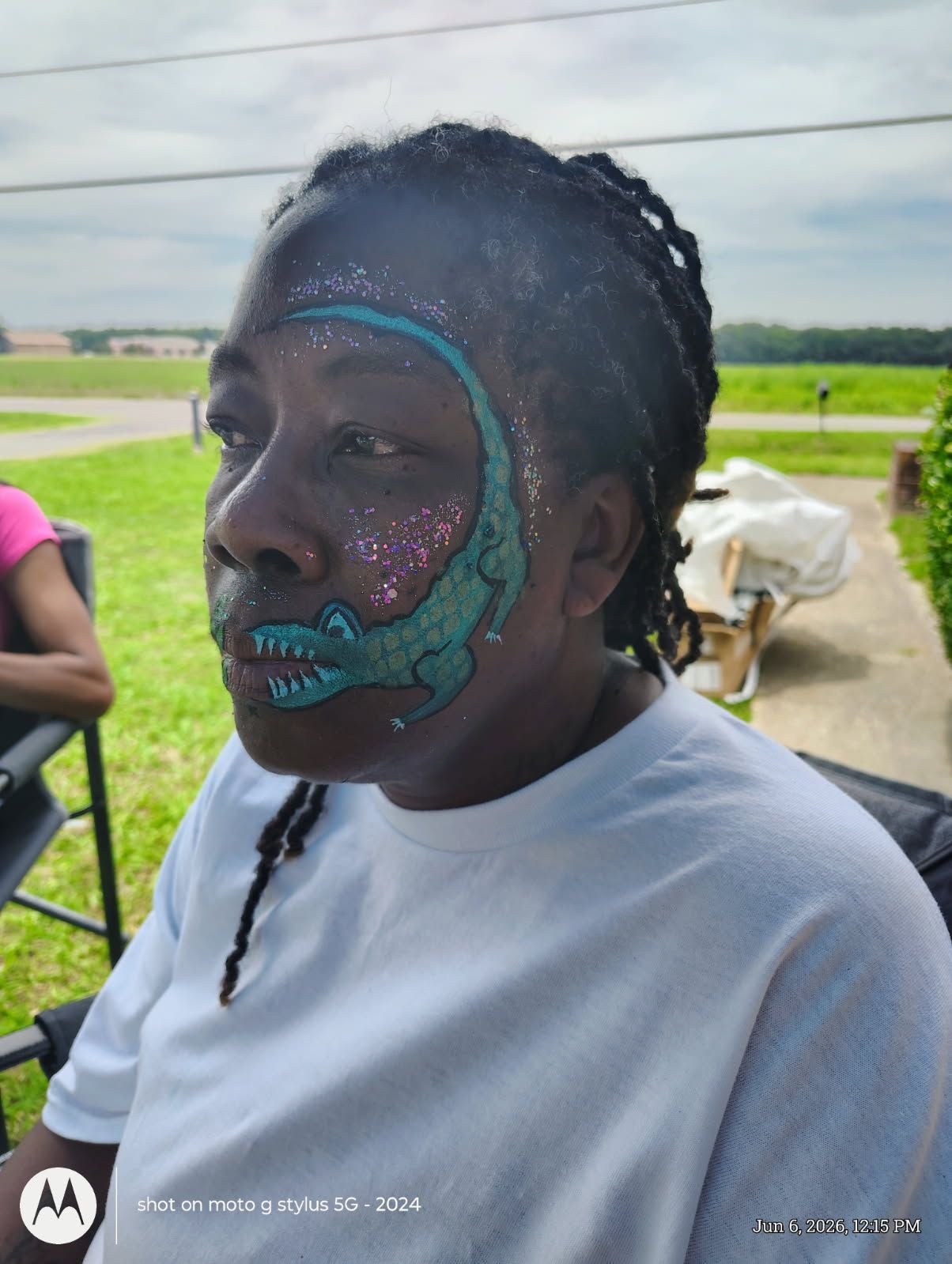 Person with teal face paint sitting outdoors in a field, wearing a white shirt and braids.