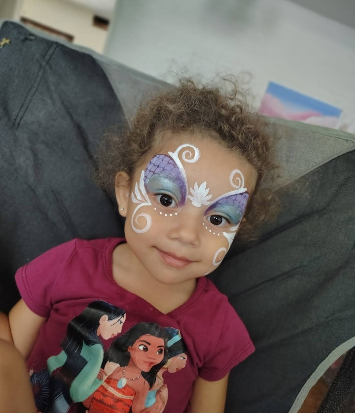 Child with butterfly face paint and a magenta shirt sits on a dark couch, looking at the camera.