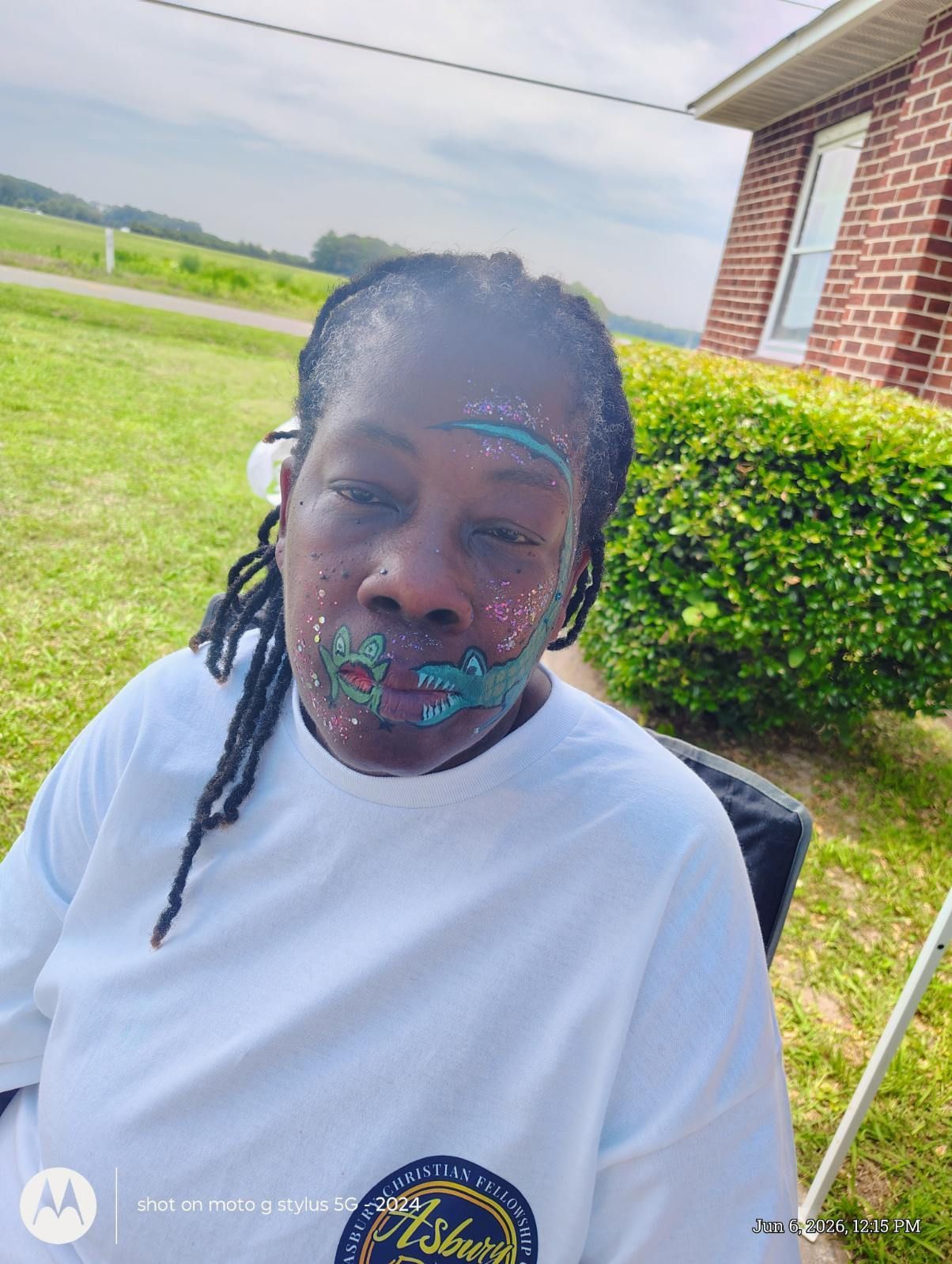 Person seated outdoors beside a brick building and hedge, wearing a white shirt and face paint.