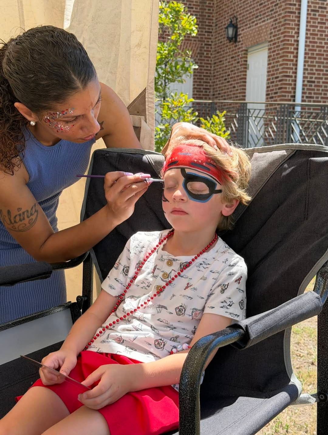 Adult applying face paint to child seated in a chair outdoors, both wearing costumes.