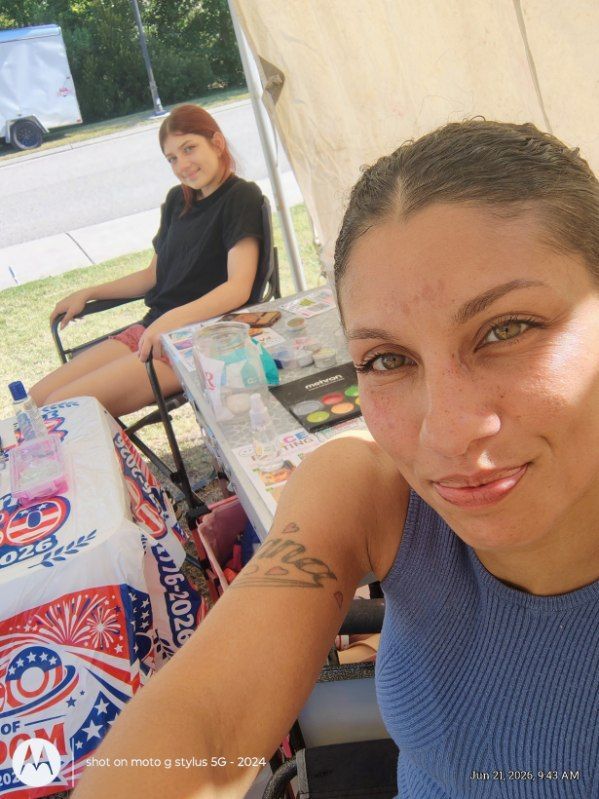 Two people at a picnic table, one taking a selfie under a tent with drinks and snacks nearby.