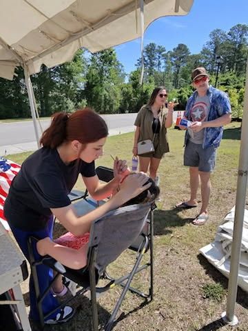 Woman at a canopy booth painting a seated child’s face while another child watches nearby.