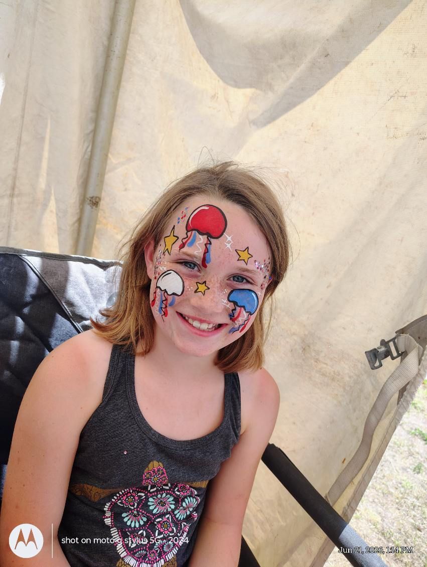 Young child with colorful face paint smiling in a tent, wearing a black tank top.