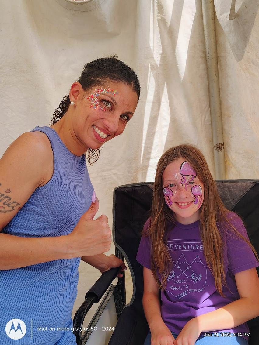 Two smiling people with face paint sit indoors, one giving a thumbs-up.