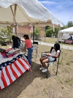 Outdoor booth under white tents with a patriotic tablecloth; people sit and stand around it.