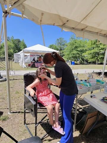 Person braids a seated child’s hair under a white canopy at an outdoor event.