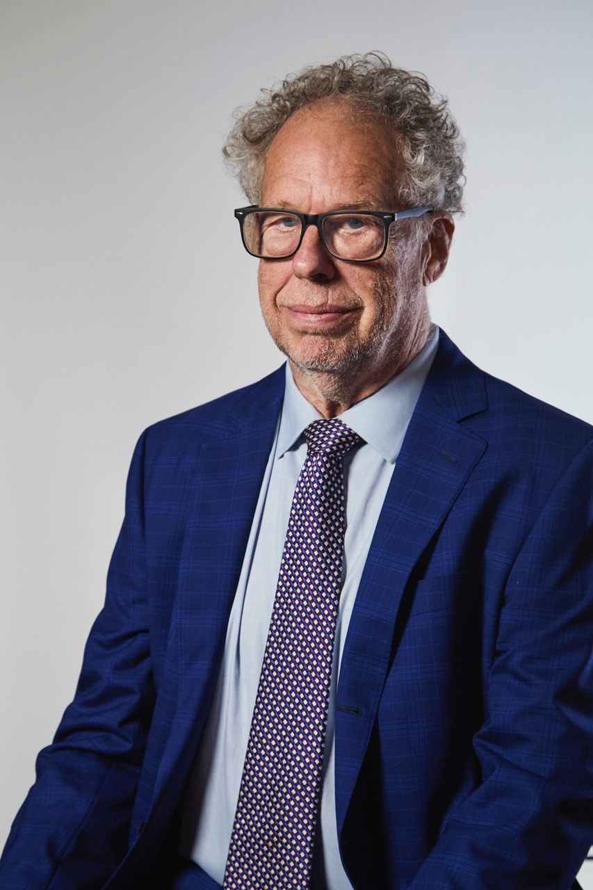 Studio portrait of a man in a blue suit, white shirt, and patterned tie, wearing glasses and smiling slightly