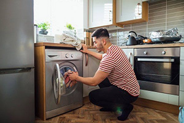 Un hombre está arrodillado en una cocina cargando ropa en una lavadora.