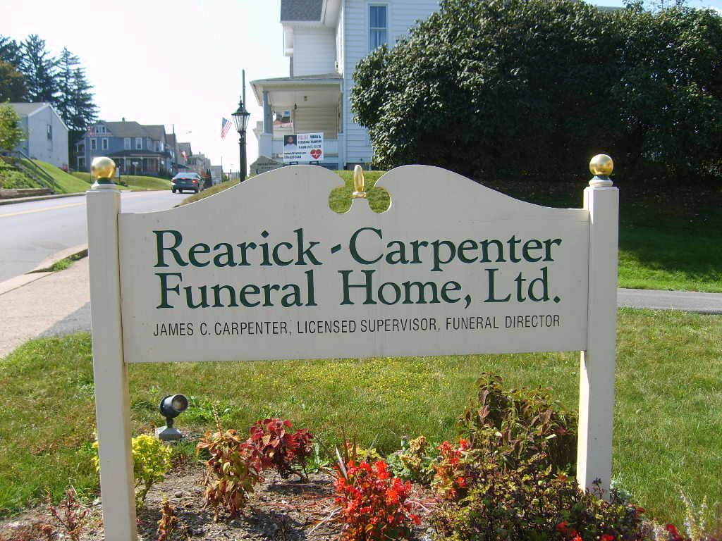 Sign for Rearick-Carpenter Funeral Home, Ltd. on a grassy lawn, with a street and buildings in the background.