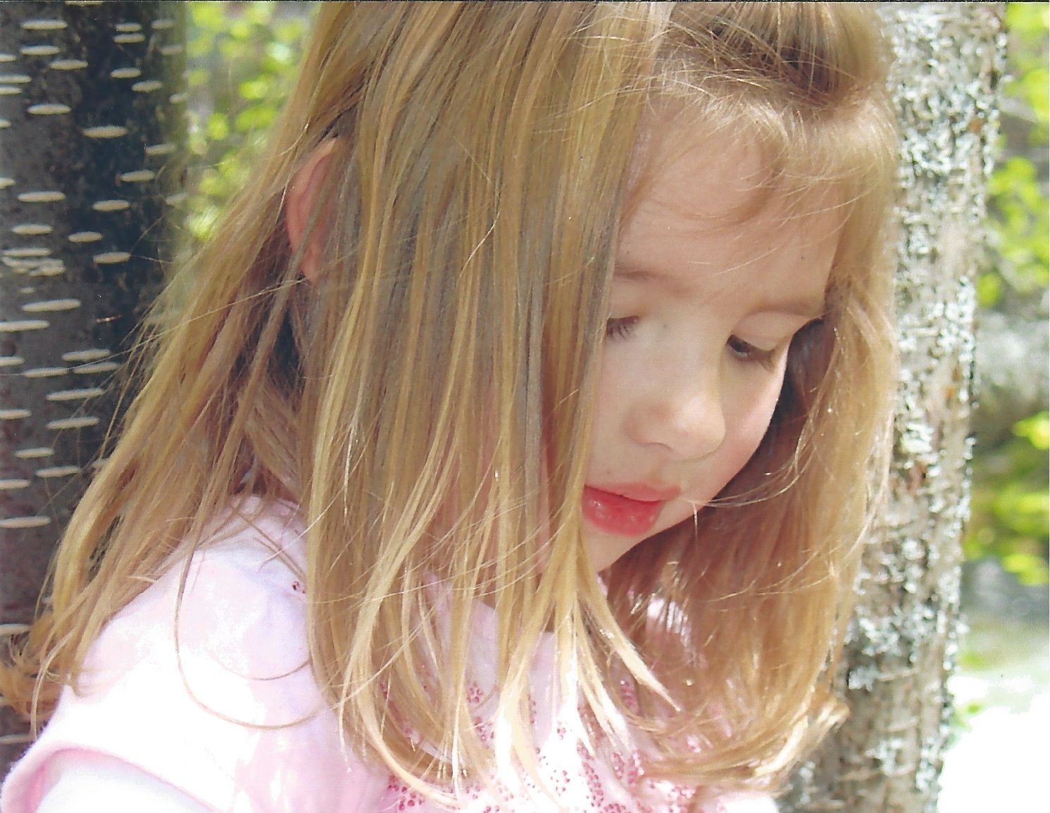 A little girl in a pink shirt is leaning against a tree