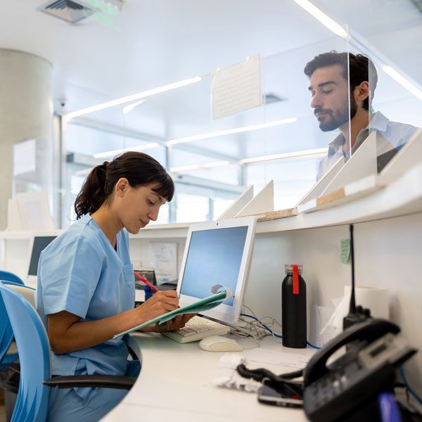 Nurse reviewing a chart at a hospital desk behind a glass partition, with a patient standing nearby.
