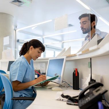 Nurse reviewing a chart at a hospital desk behind a glass partition, with a patient standing nearby.