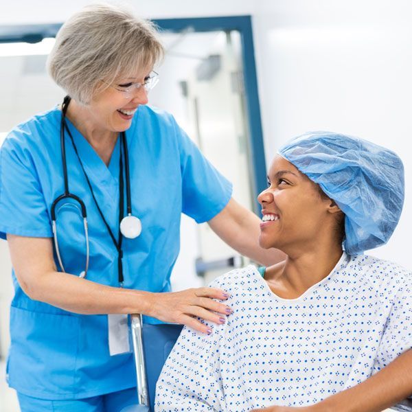 Nurse in blue scrubs smiling with a patient in a hospital gown and cap at bedside