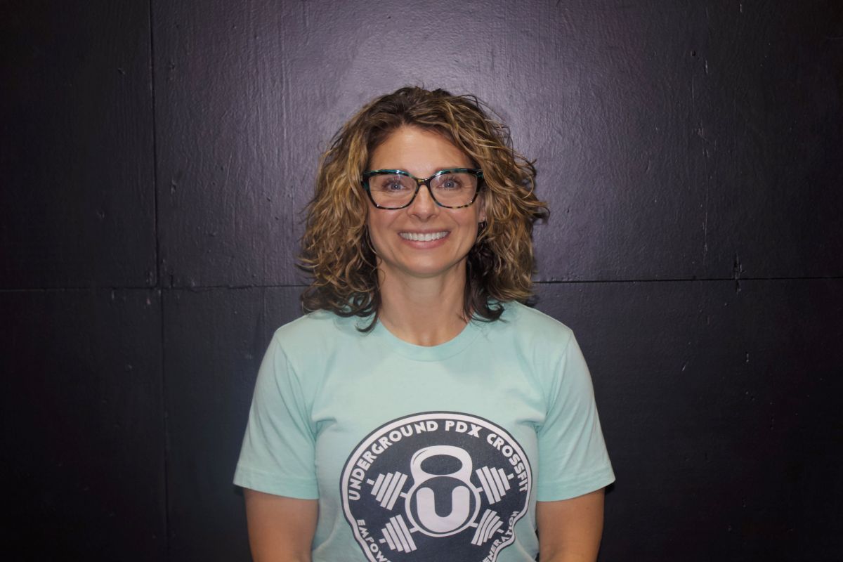 Woman with curly blonde hair smiles, wearing a yellow floral top, against a dark, industrial background.