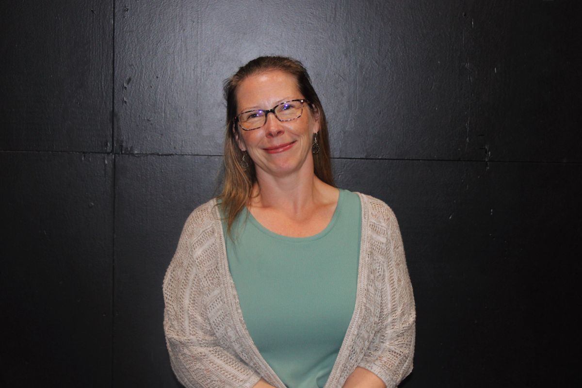 Woman with glasses smiles, in front of a dim basement gym background. She wears a black cardigan and a silver cross necklace.