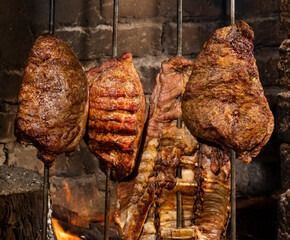 Tres trozos de carne asándose en asadores verticales sobre fuego abierto.