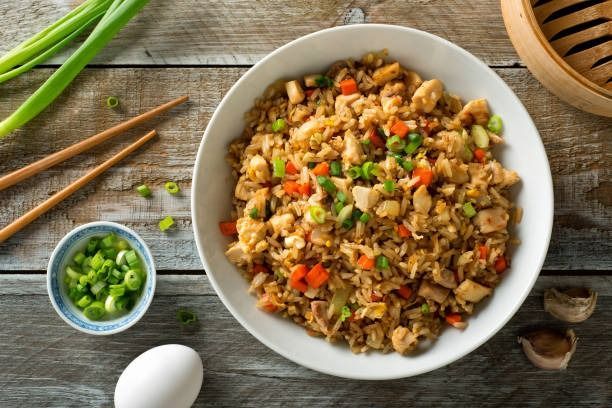 Un bol de arroz frito con verduras y cebolletas sobre una mesa de madera, con palillos y un huevo cascado cerca.